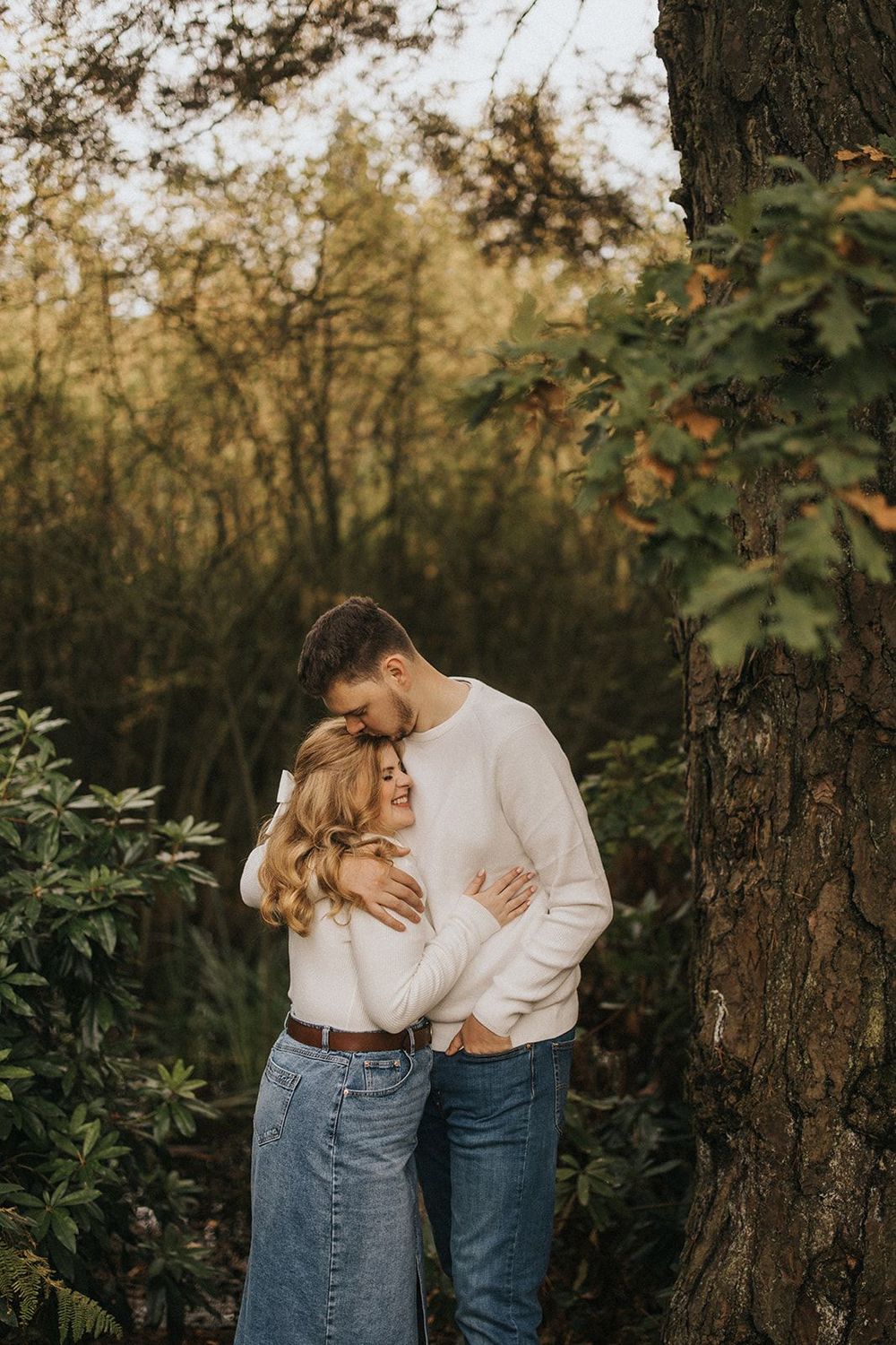 A newly engaged couple embracing one another near a large tree trunk in an autumn forest setting. Captured by Lincolnshire photographers.