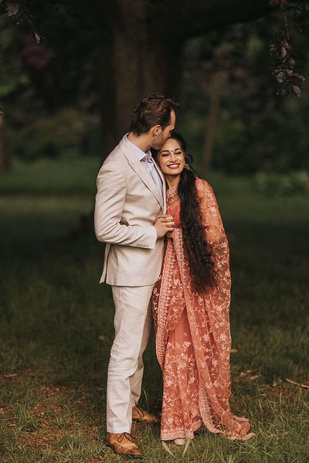 A romantic couple in formal attire walks through a grassy meadow during sunset, with her wearing a peach-colored dress.
