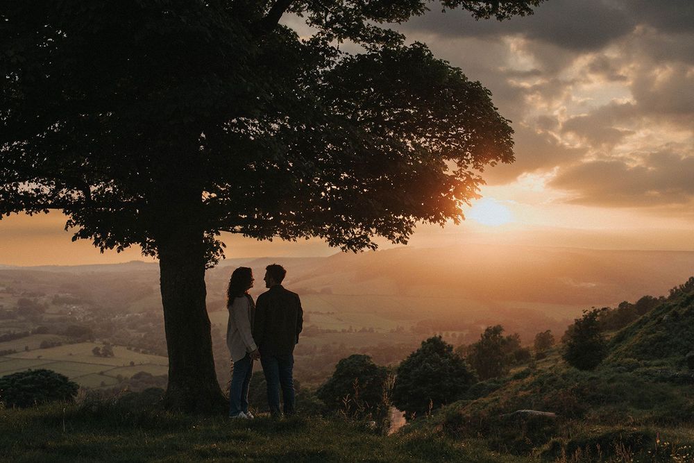 A couple stands in silhouette against a dramatic sunset sky beneath a large tree on a hilltop. Captured by Lincolnshire photographers.