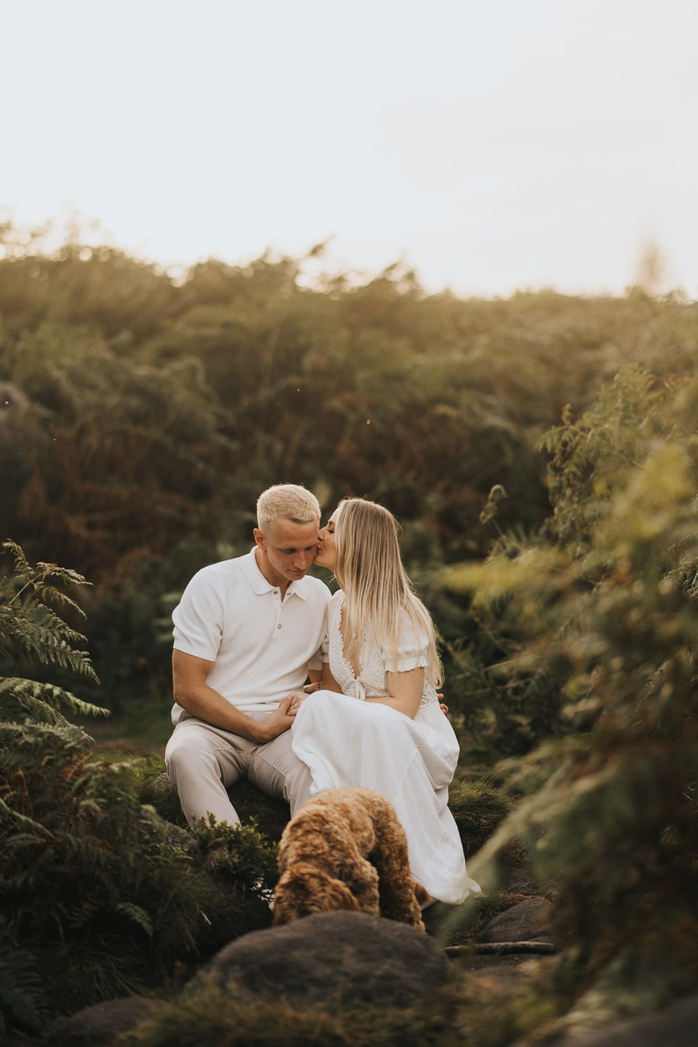 A couple in white clothing shares an intimate moment among ferns and greenery during golden hour.