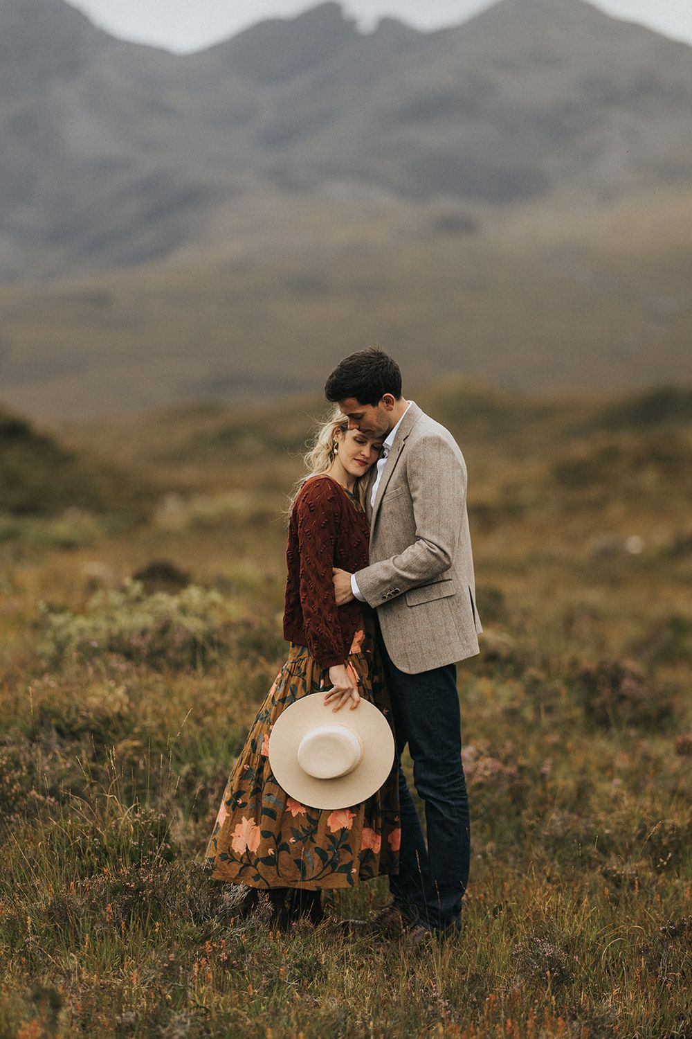 A romantic couple embraces in a rustic mountain landscape with moody skies and natural terrain in the background.