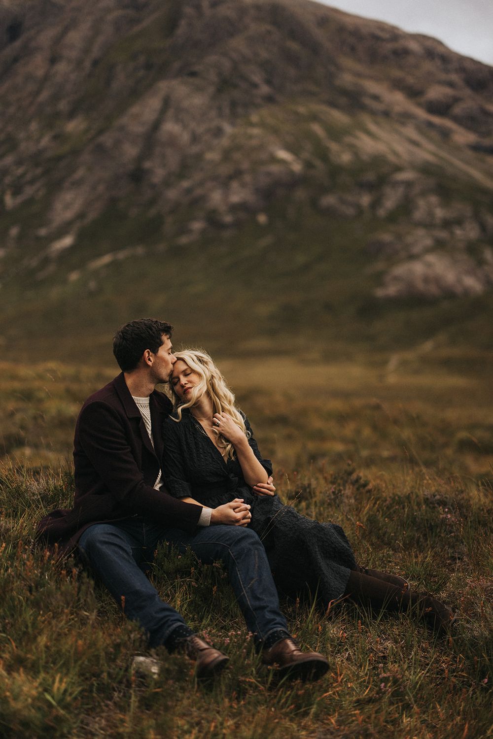 A couple shares an intimate moment while sitting together on a grassy hillside with mountains in the background.