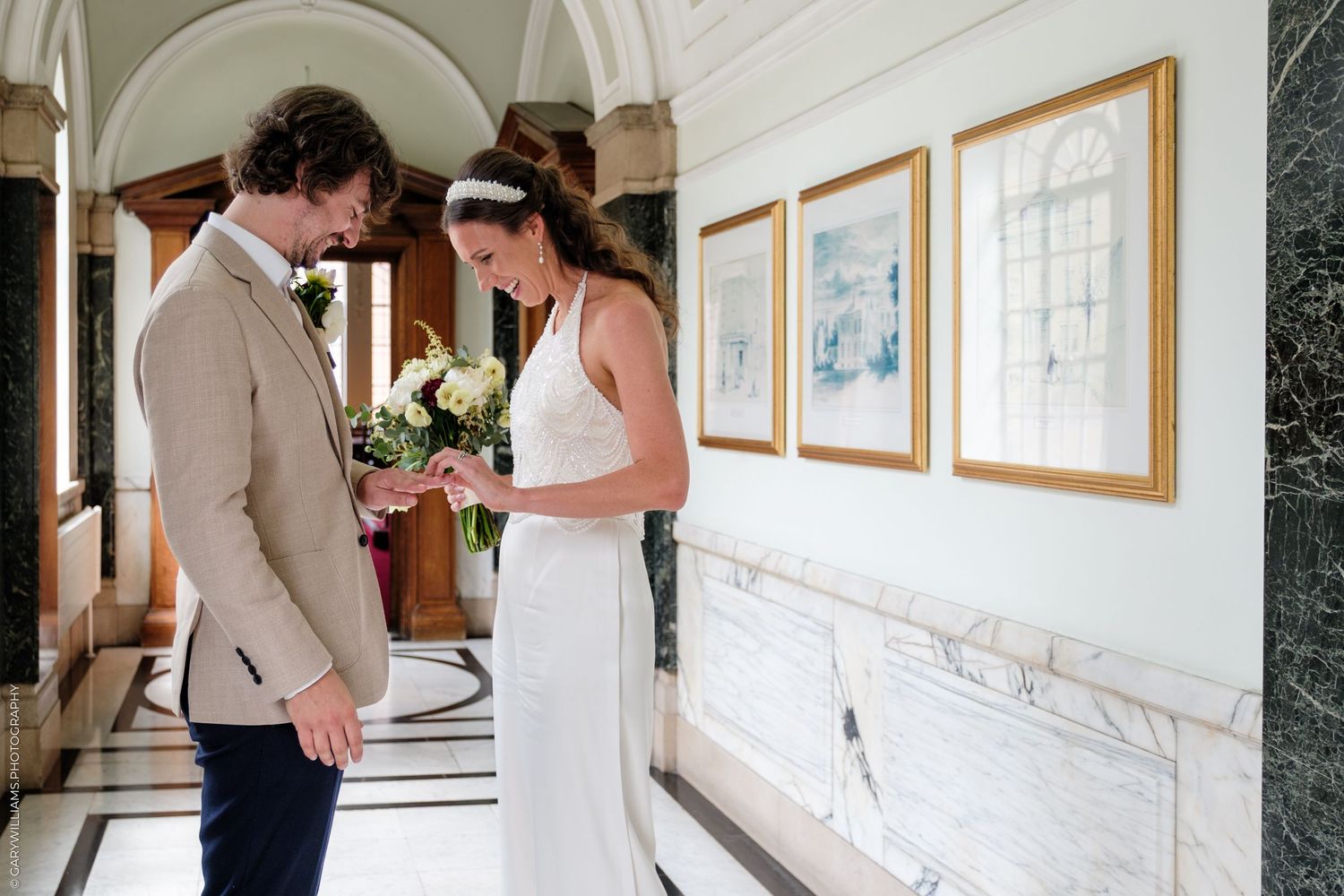 A couple shares an intimate first look moment in a hallway decorated with framed artwork and white columns.