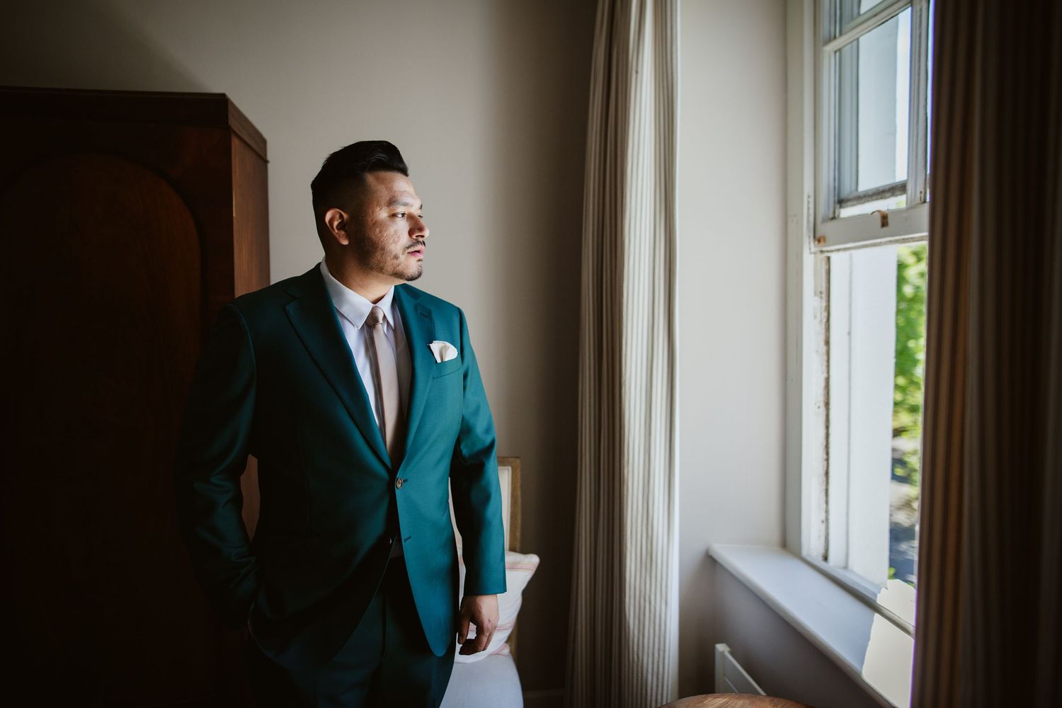 A person in a teal suit jacket stands near a window, gazing thoughtfully at natural light streaming in.
