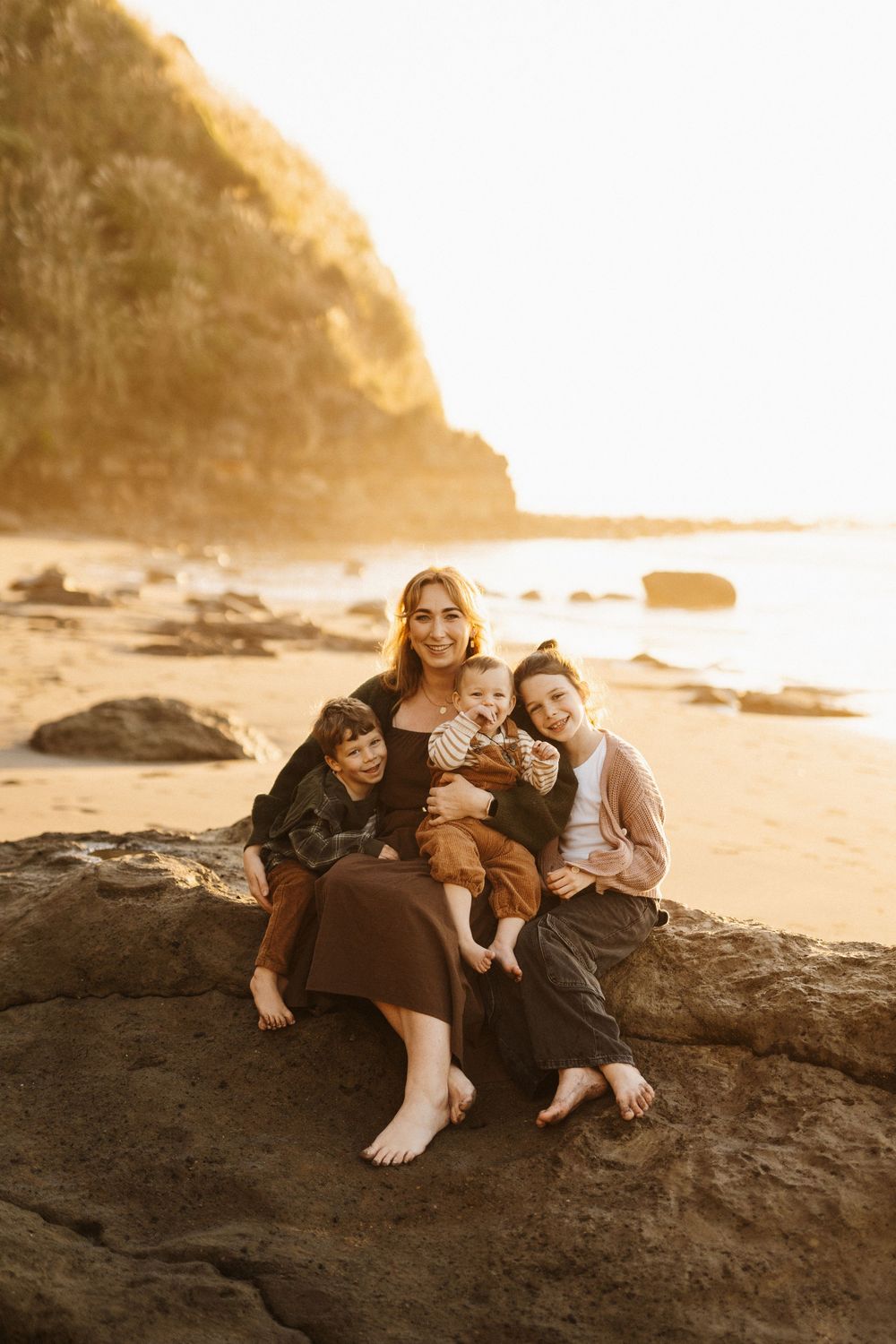 Family sitting together on coastal rocks at sunset with golden light and cliffside in background at the beach.