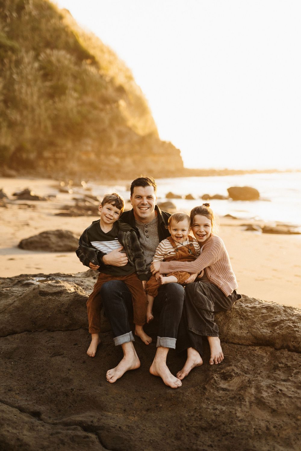 A family sits barefoot together on coastal rocks at sunset with cliffs and ocean in the background.