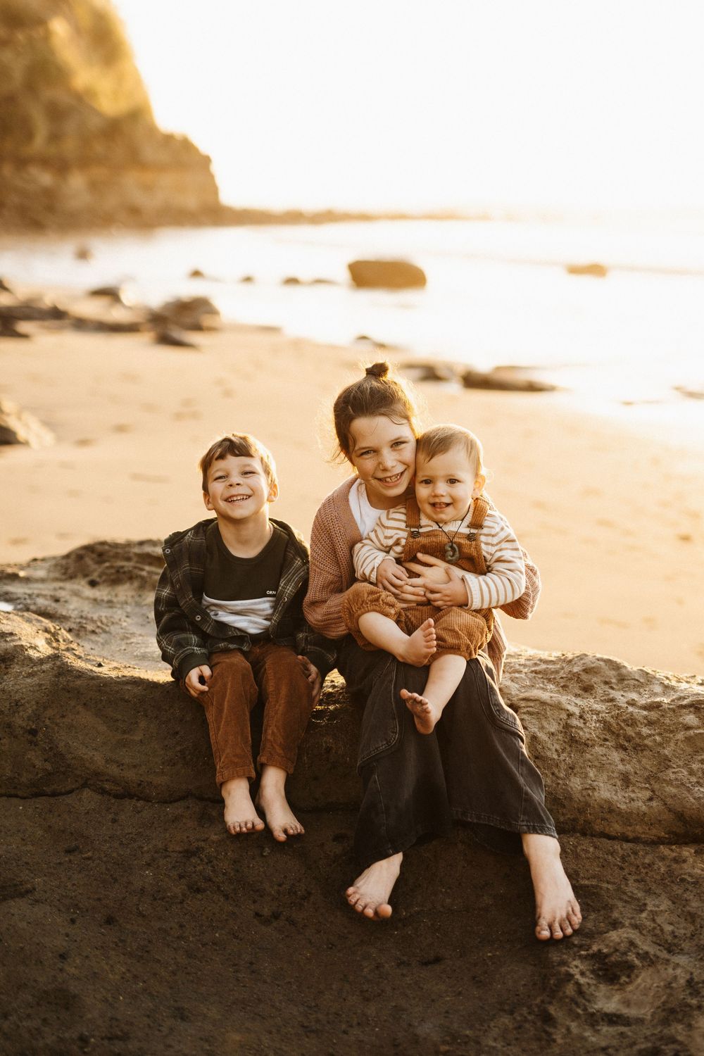 Three people sit together on coastal rocks at sunset, barefoot and smiling at a golden-lit beach.