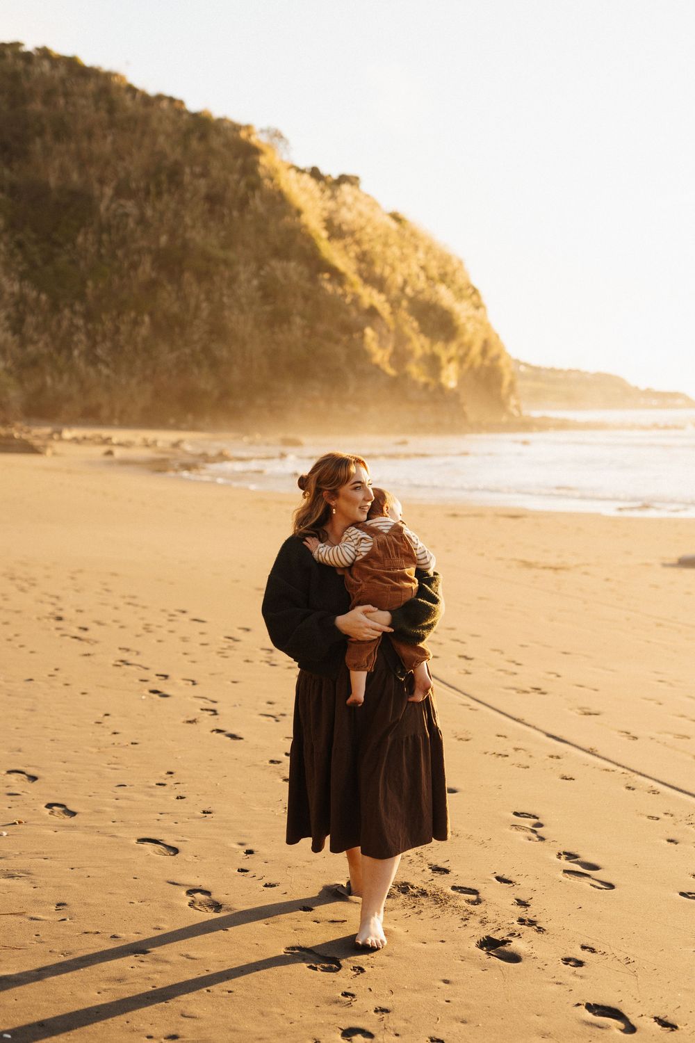 A figure in a long dress stands on a golden beach at sunset holding a small bundle, with cliffs in the background.