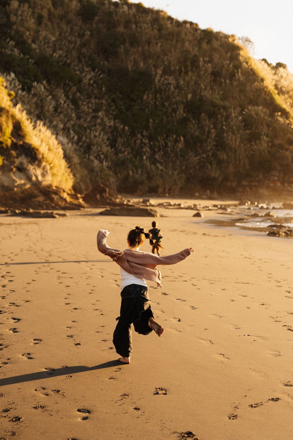 A person joyfully leaps in the air on a golden sandy beach with rugged coastal cliffs behind them.