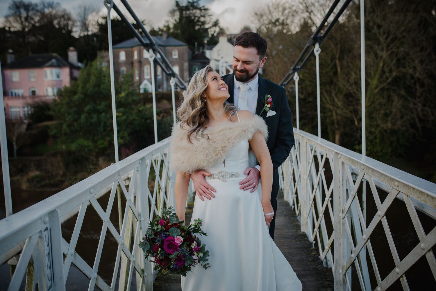 Happy couple embraces on white wooden bridge decorated with floral arrangements during outdoor wedding celebration.