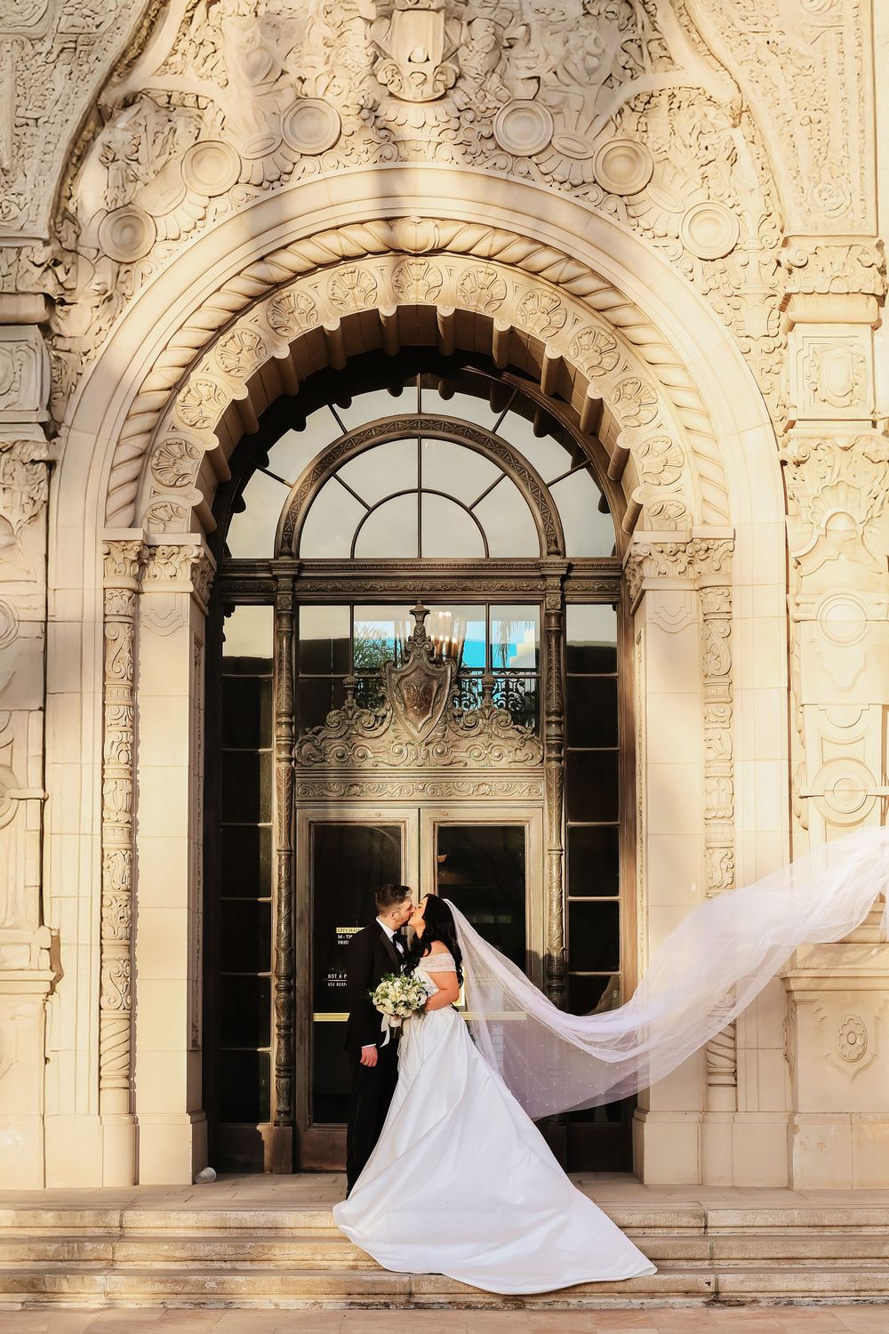 A couple embraces in front of an ornate stone Beverly City Hall archway with a flowing white wedding veil caught in the breeze.