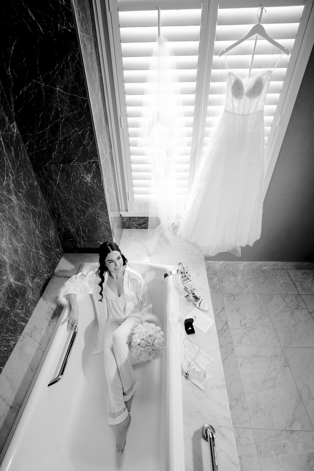 A white wedding dress hangs by a window with white shutters while preparations are laid out in a marble bathtub.