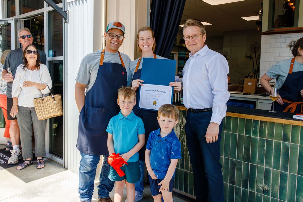 A small business owners and children stand proudly displaying a certificate outside their shop entrance.