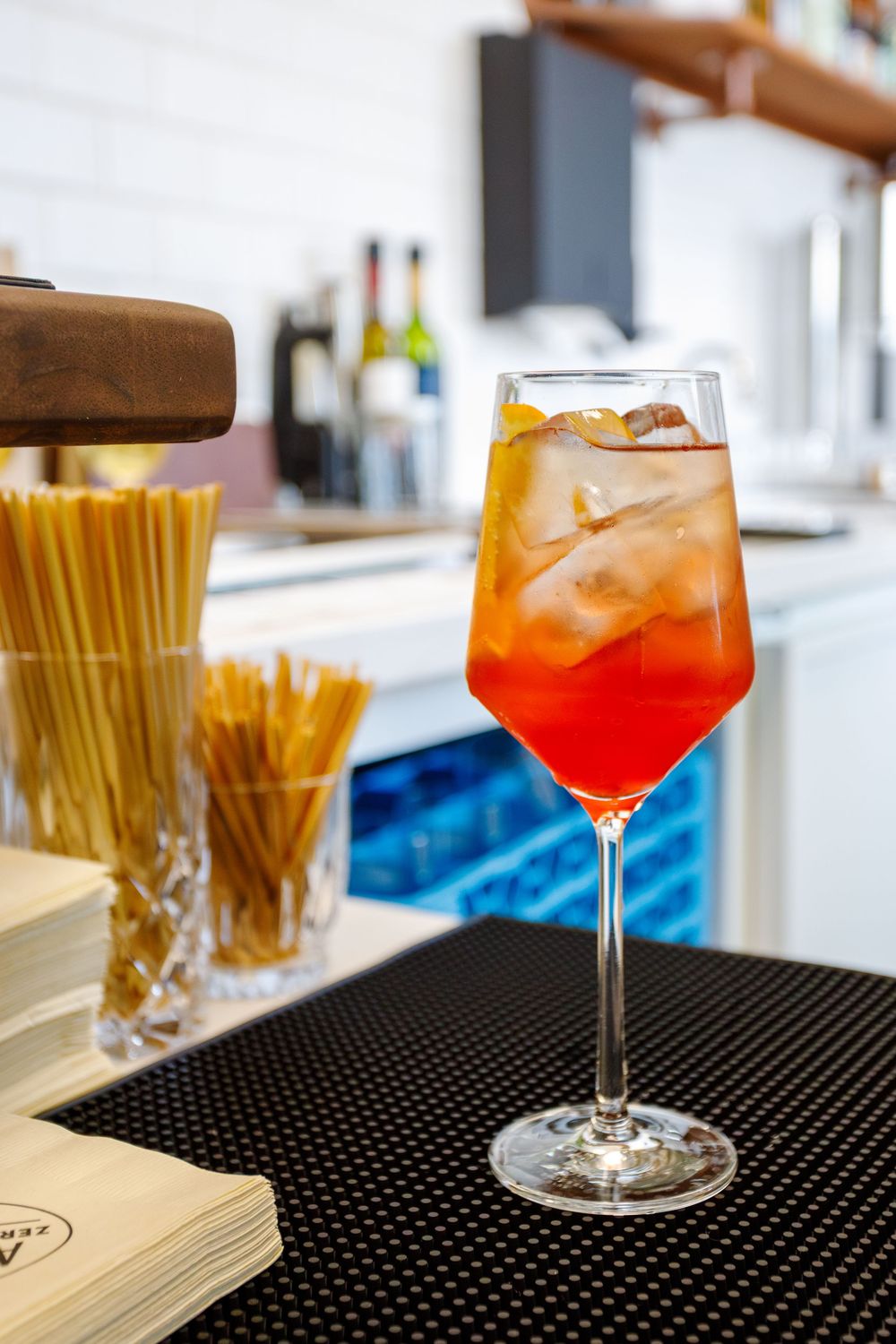 An orange-red cocktail with ice served in a wine glass on a black bar mat next to jars of straws.
