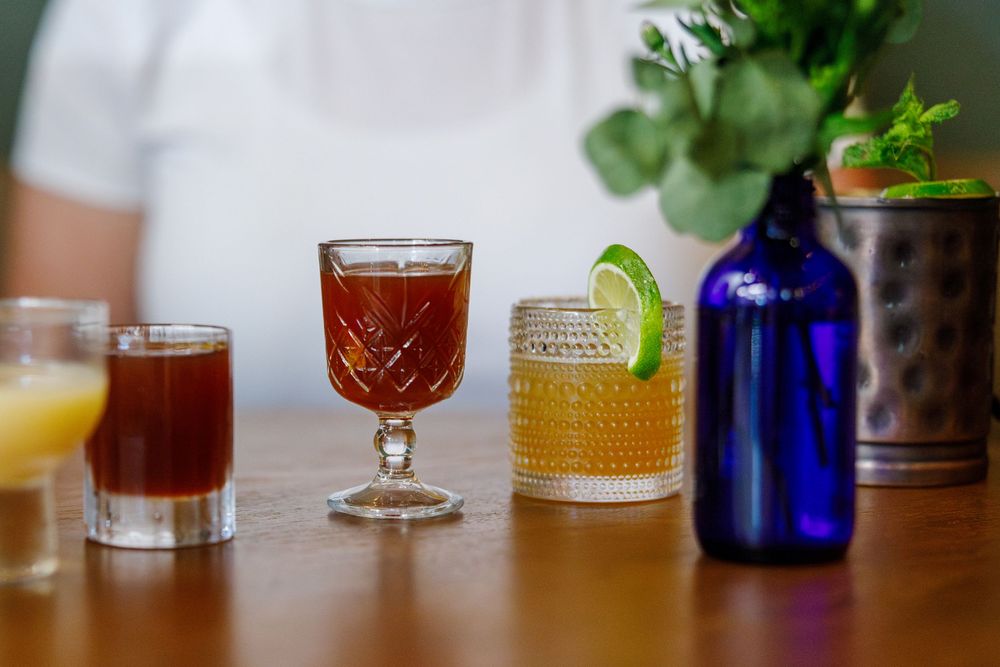 Variety of colorful craft cocktails in vintage glassware arranged on wooden table with lime garnish and blue bottle.