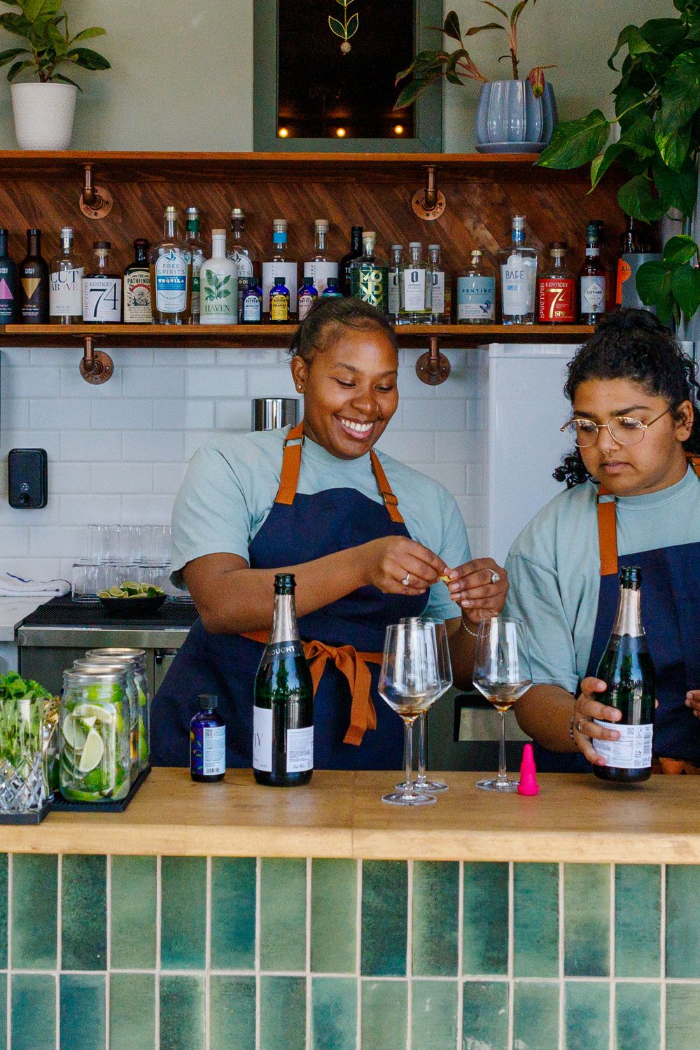 Two bartenders in blue aprons work behind a counter with bottles of spirits and cocktail ingredients at a modern bar setting.