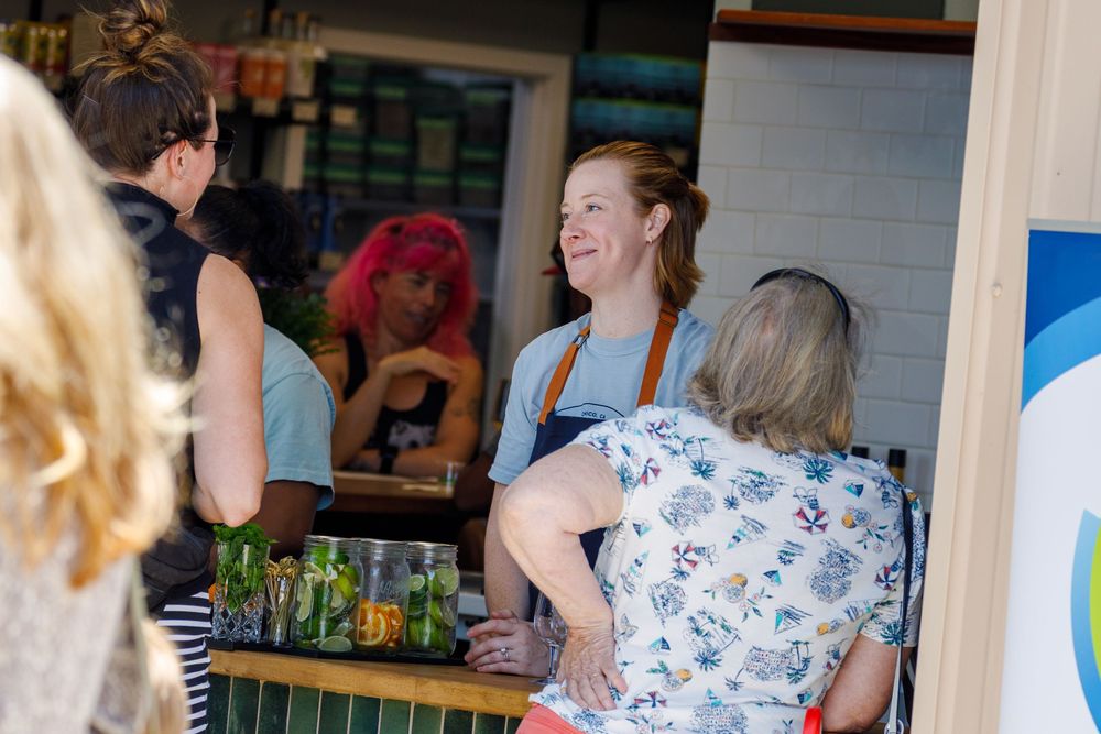 A server wearing an apron chats with customers at a casual bar counter with bottled drinks displayed.