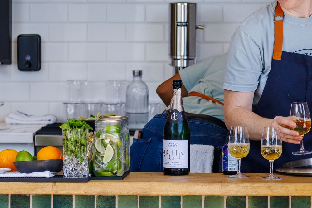 A bartender pours drinks at a counter with fresh citrus fruits and herbs in glass jars on display.