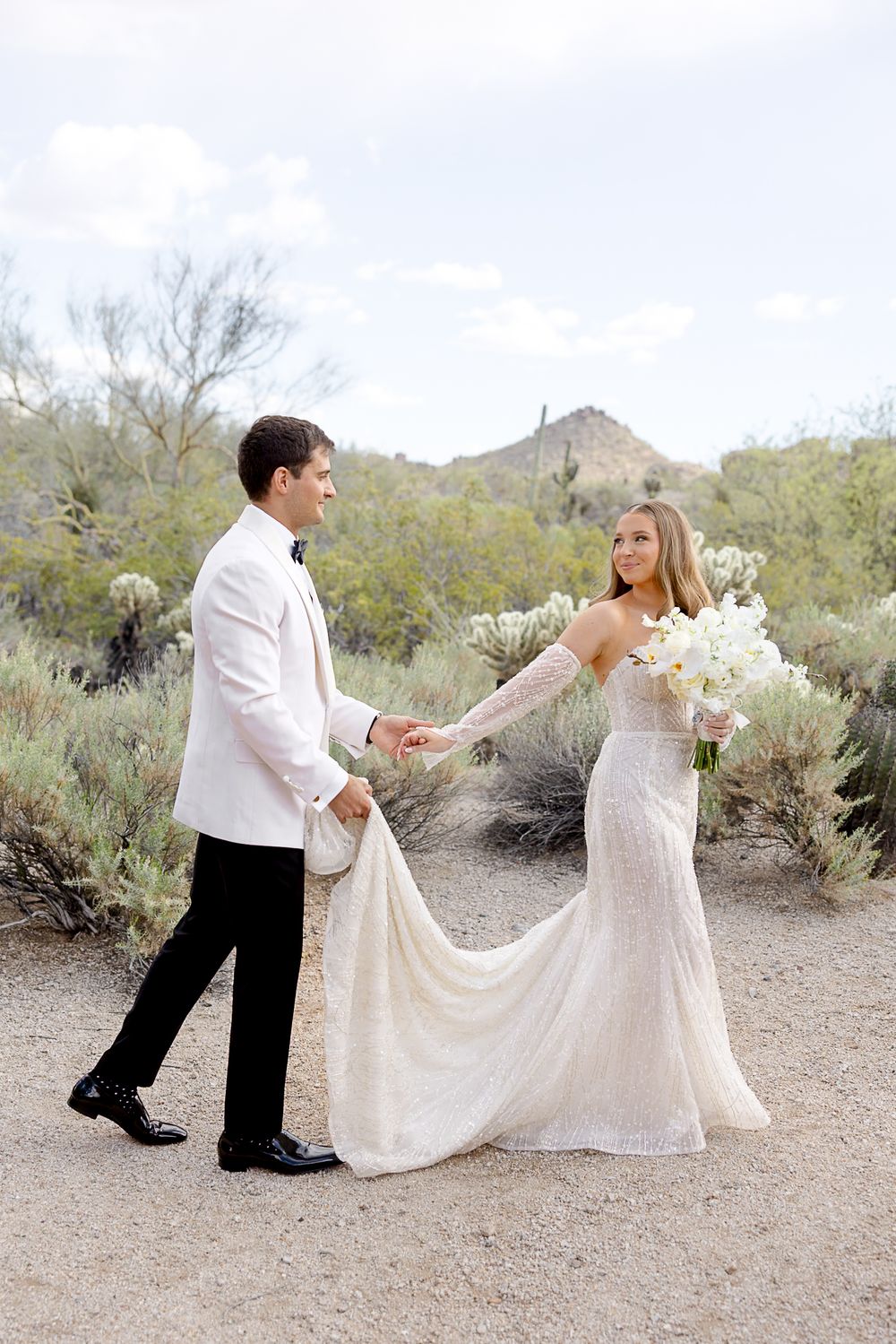 A romantic desert scene shows a couple in wedding attire playfully dancing with the bride's dress flowing in the outdoor setting.