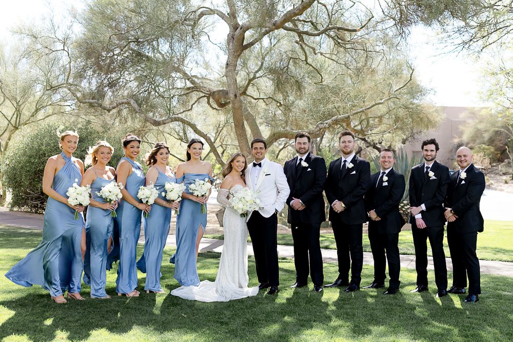 A wedding party wearing black suits and light blue dresses poses together under a large tree in a park setting.