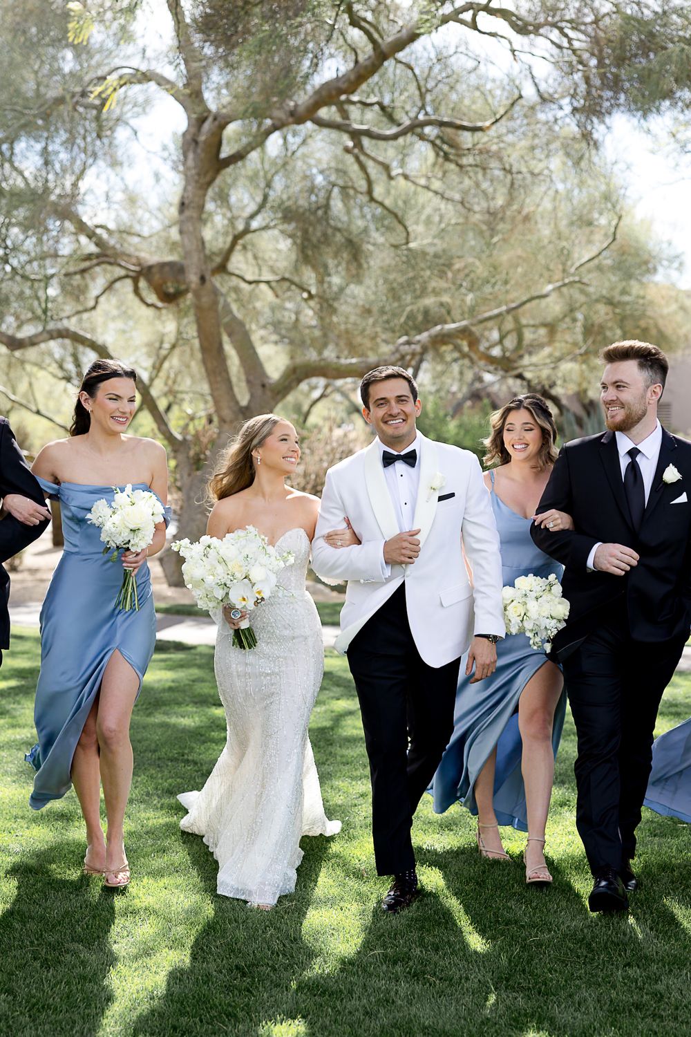 A wedding party walks and laughs together on a sunny day under olive trees in a garden setting.
