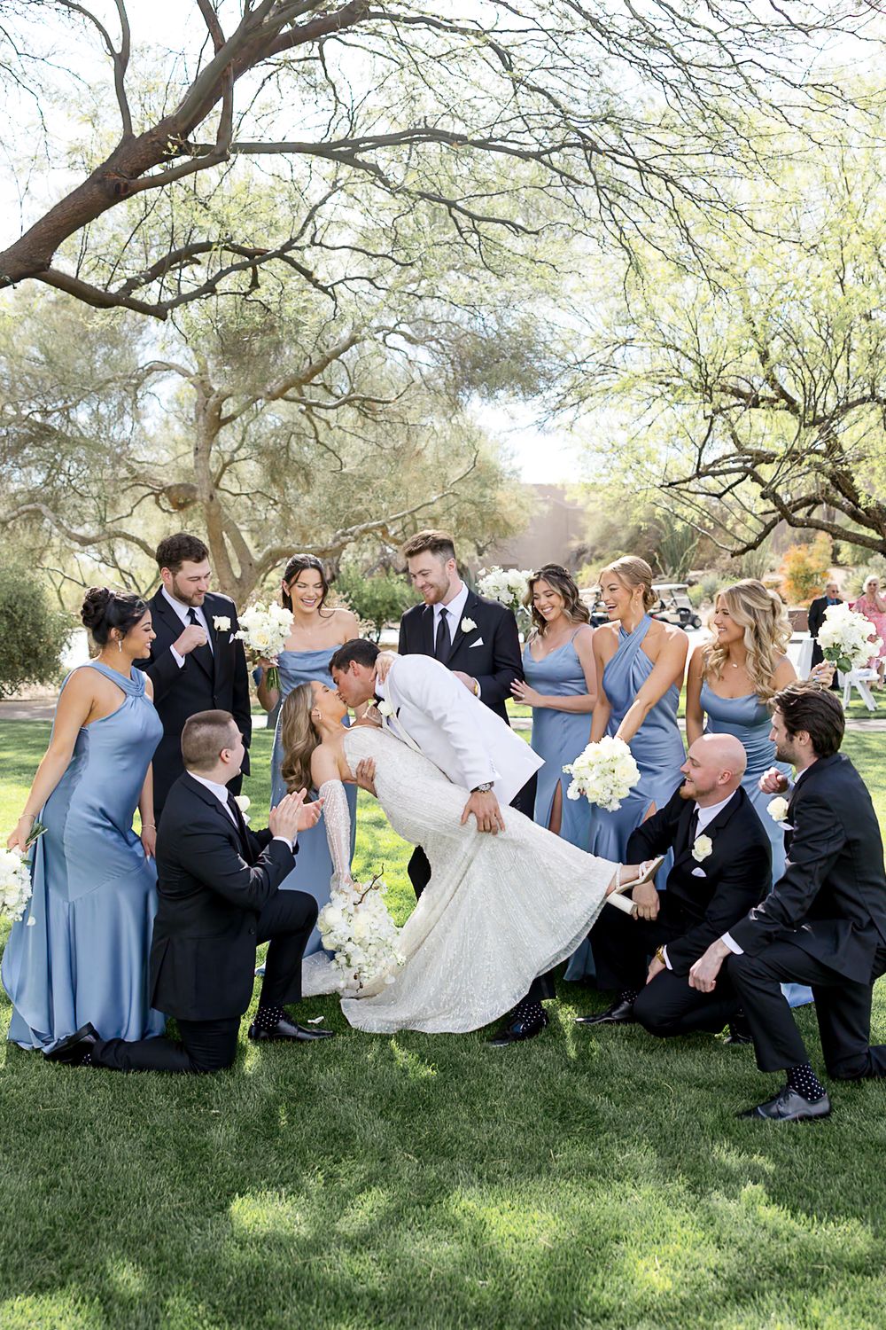 Wedding party poses on grass under trees at outdoor ceremony with bridesmaids in blue dresses and groomsmen in dark suits.