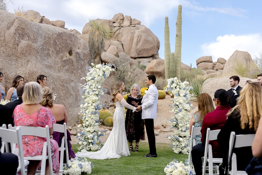A desert wedding ceremony with guests seated among giant boulders and saguaro cacti surrounded by white floral arrangements.