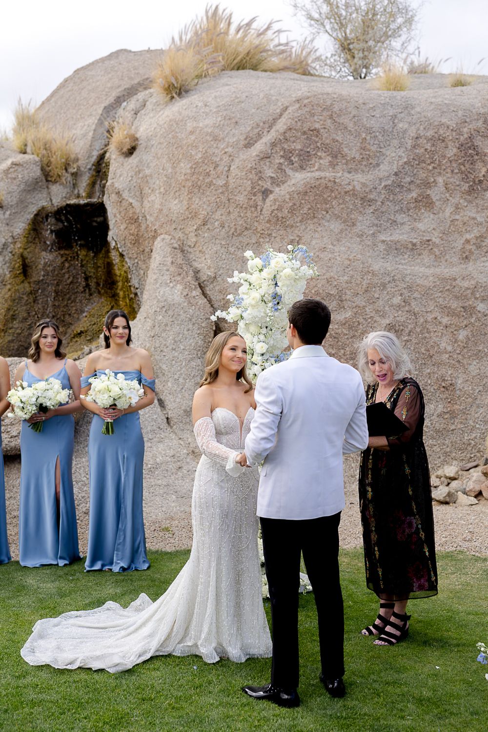 An outdoor wedding ceremony takes place in front of large desert rock formations with bridesmaids in blue dresses.