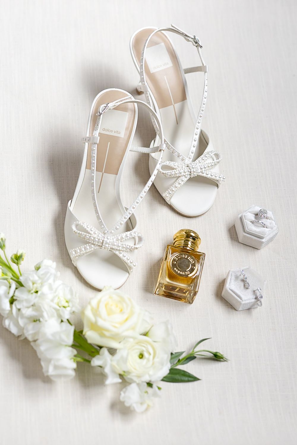Elegant white wedding accessories including high heel sandals, perfume bottle, and white roses arranged on light background.