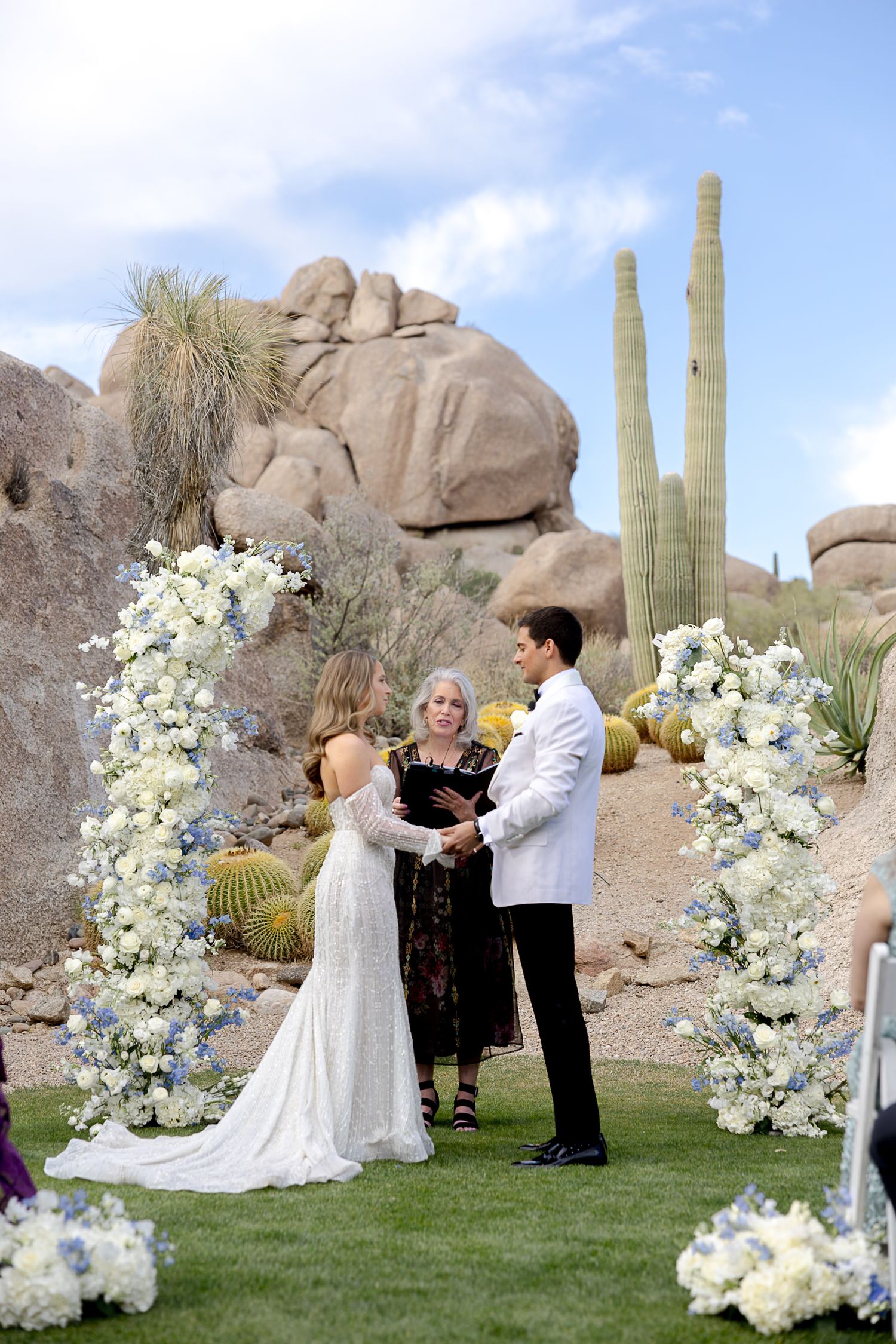 A desert wedding ceremony unfolds beneath a circular floral arch with white blooms and cacti in the background.
