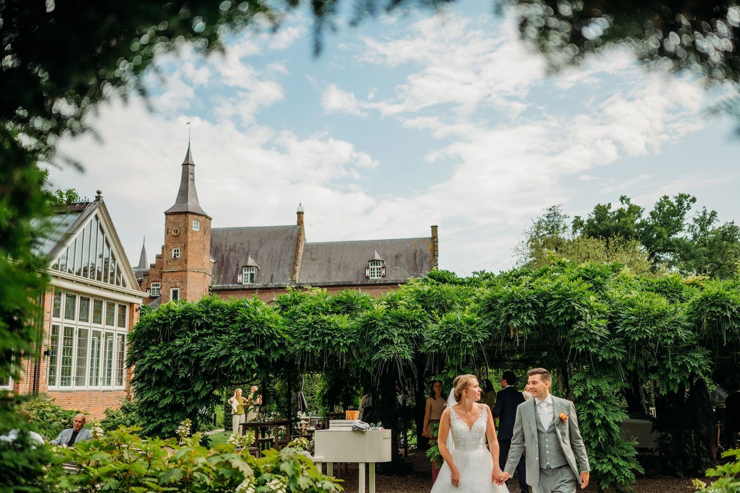 A couple walks through a lush garden path with a gothic-style building and greenhouse in the background on a sunny day.