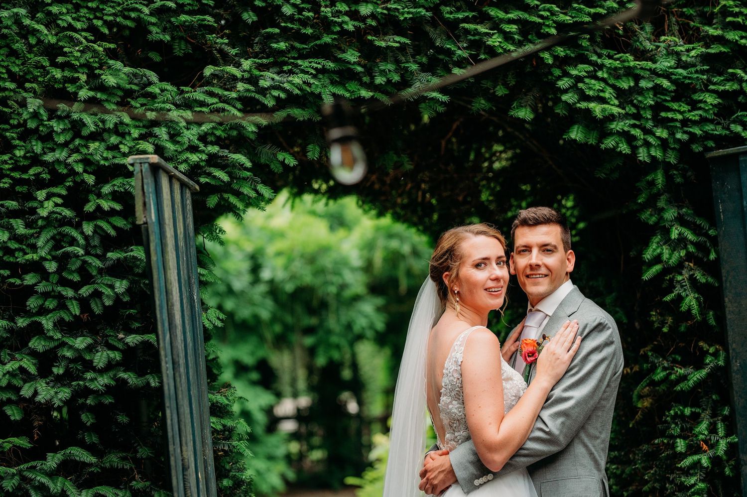 A newlywed couple poses under an ivy-covered archway in a lush garden setting during their wedding celebration.