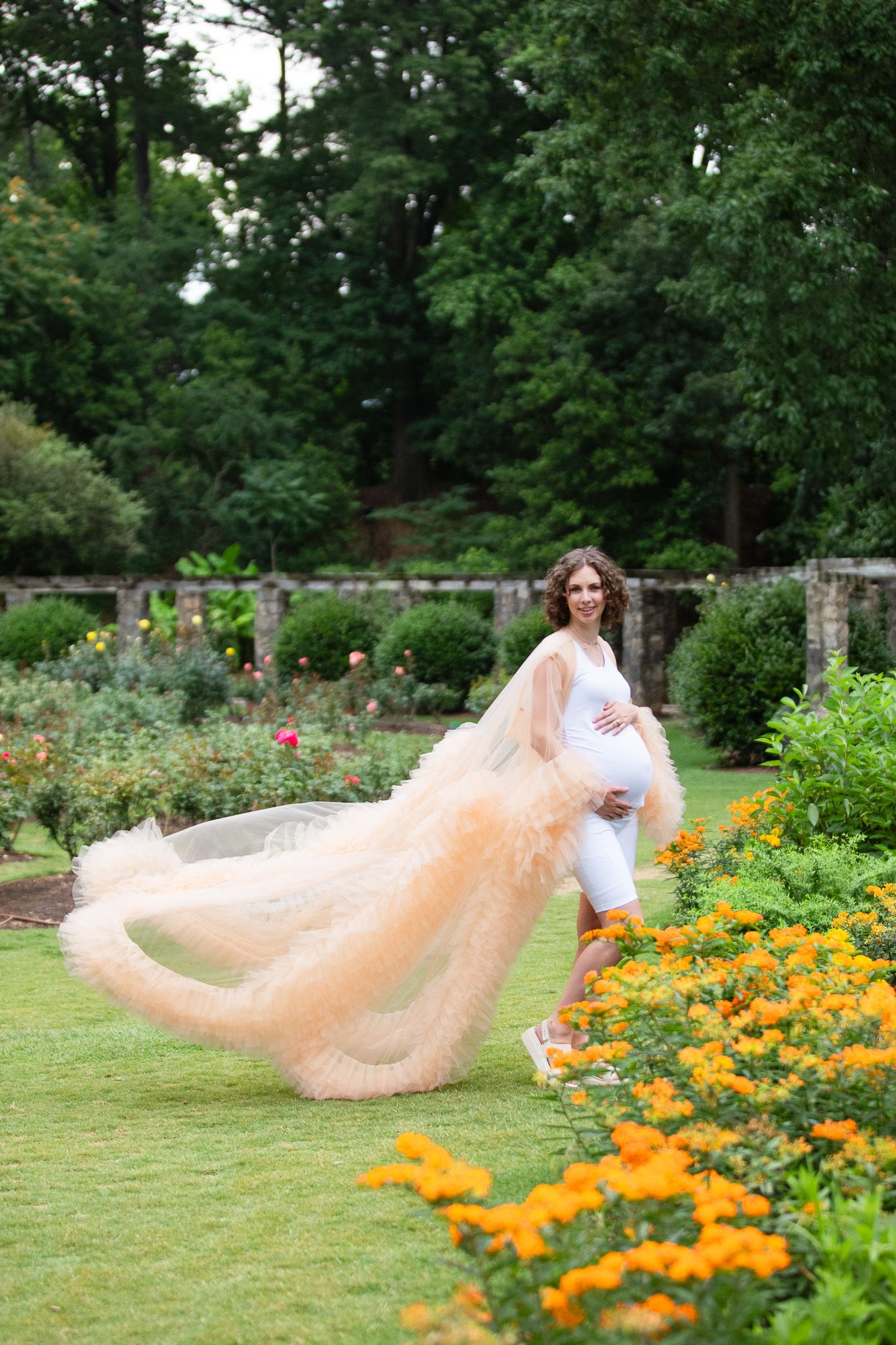 A flowing tulle gown creates ethereal movement in a garden filled with vibrant orange marigolds and lush greenery at the Raleigh Rose Garden