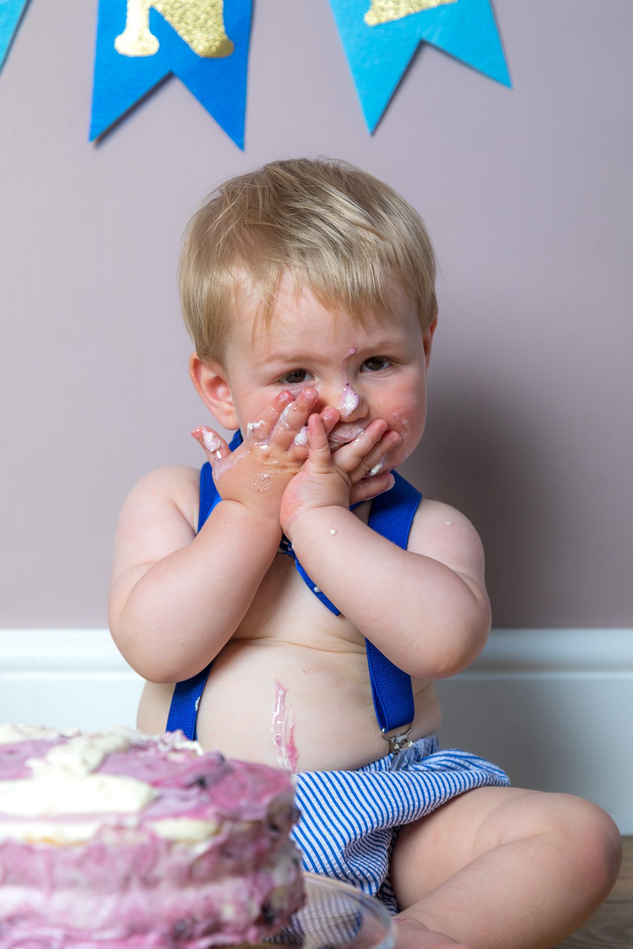 Small child in blue overalls giggles while sitting next to a pink and white birthday cake.