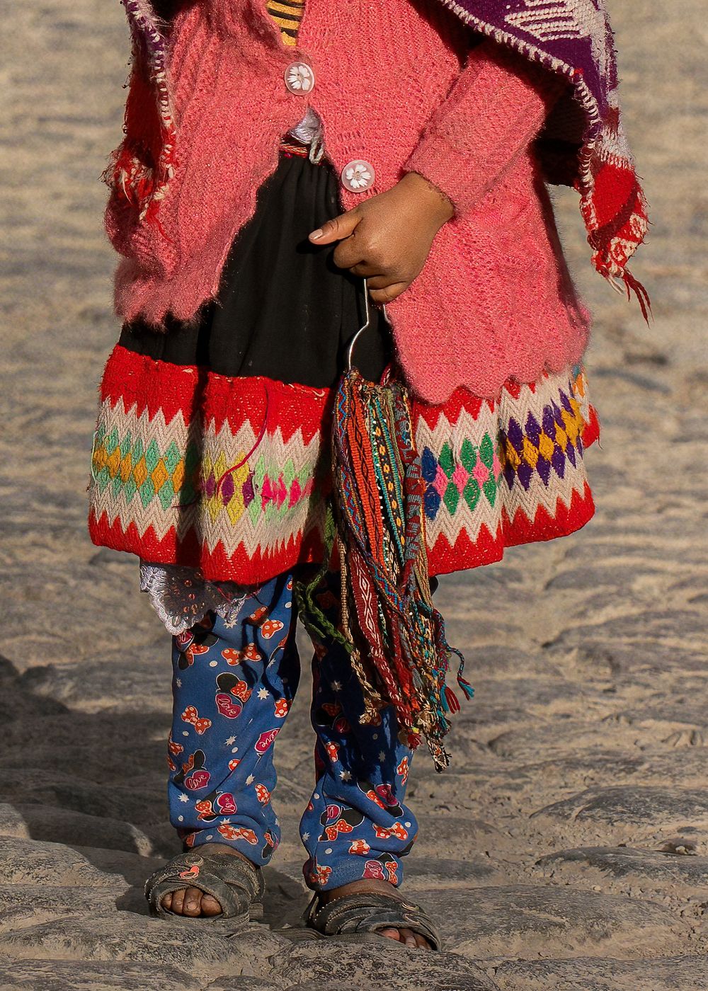 Person wearing colorful traditional clothing with pink sweater and patterned pants standing on sandy ground.