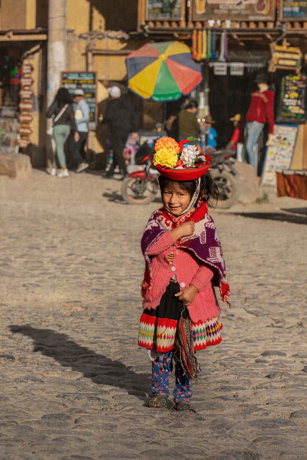 Young person in colorful traditional Peruvian clothing with a red hat stands on cobblestone street in Peru.