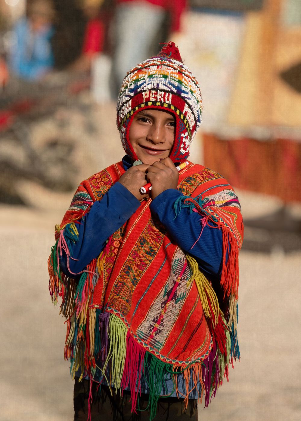 A person wearing a colorful traditional Peruvian poncho and hat with tassels stands against a blurred outdoor background.