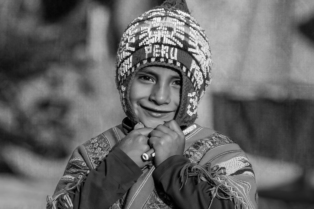 Black and white portrait of someone wearing a traditional Peruvian knit hat with decorative patterns, smiling warmly.