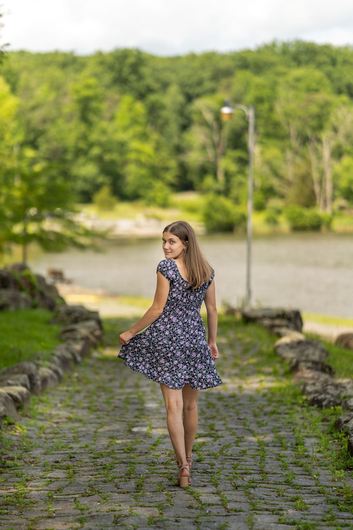 Female in a floral summer dress twirls on a cobblestone path surrounded by green trees and waterfront scenery.