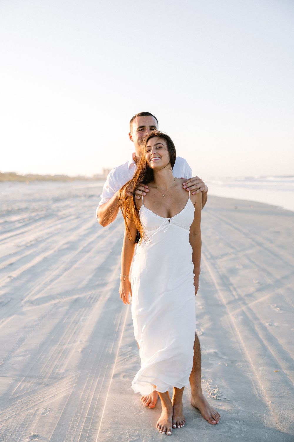 Couple in white clothing walking together on sandy beach during golden hour.