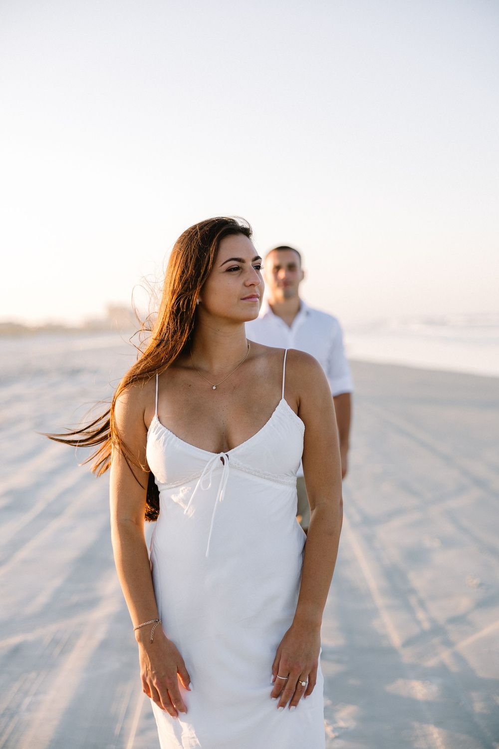 Woman in white dress poses on beach with companion in background during sunset.