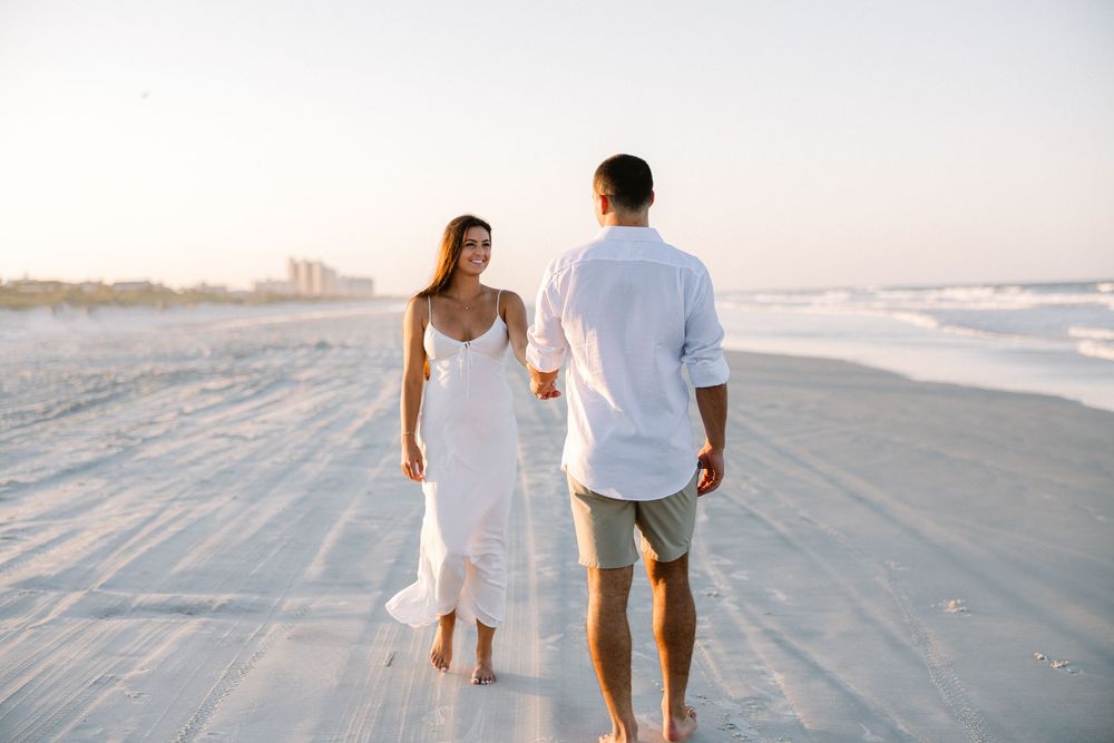 Couple walking barefoot on sandy beach at sunset wearing white and casual attire.