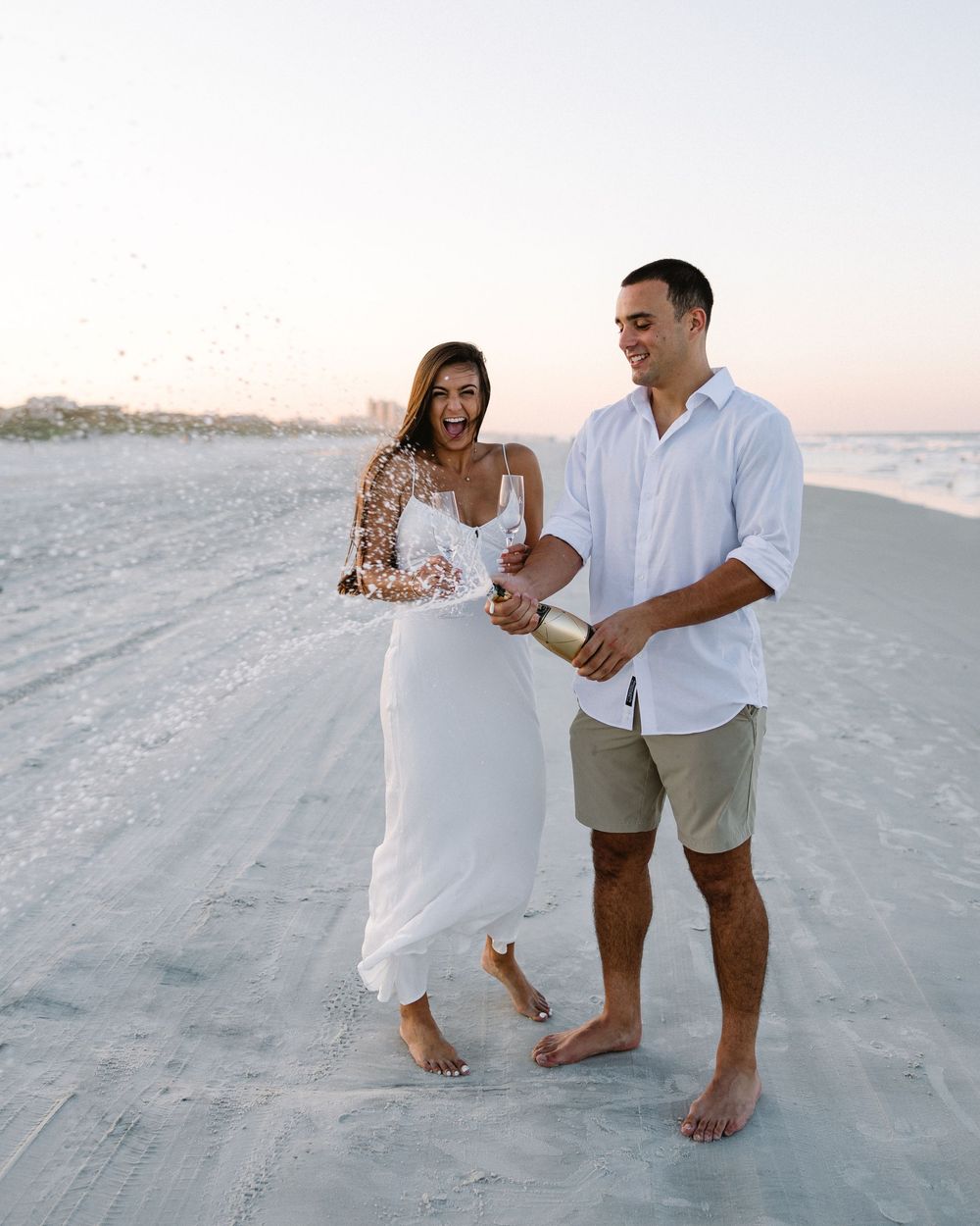 Couple popping champagne together on beach in white summer clothing.