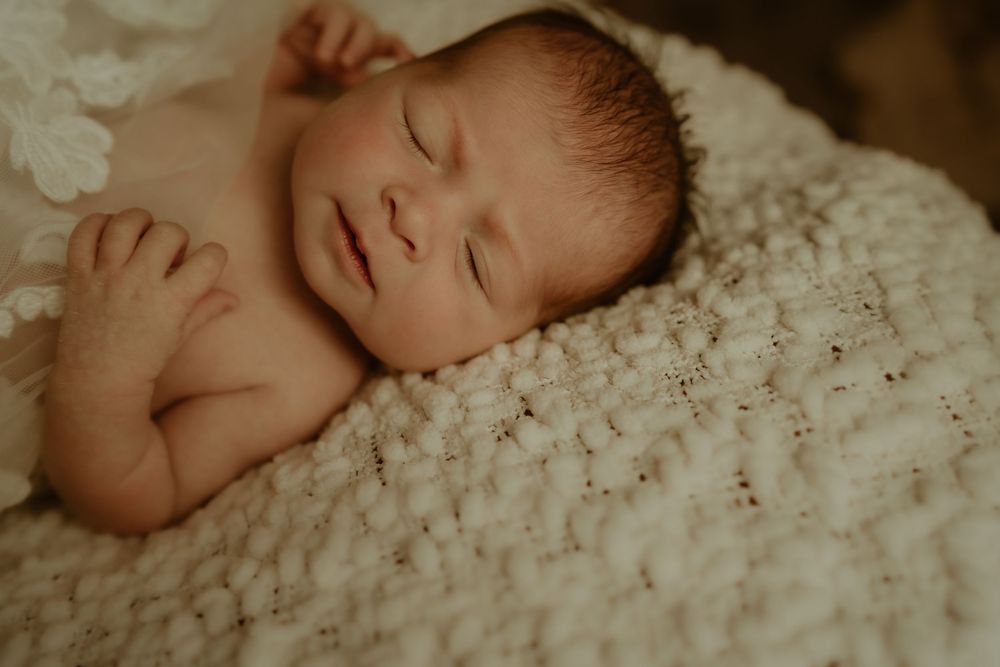 A newborn baby sleeps peacefully on a textured white blanket in warm lighting.