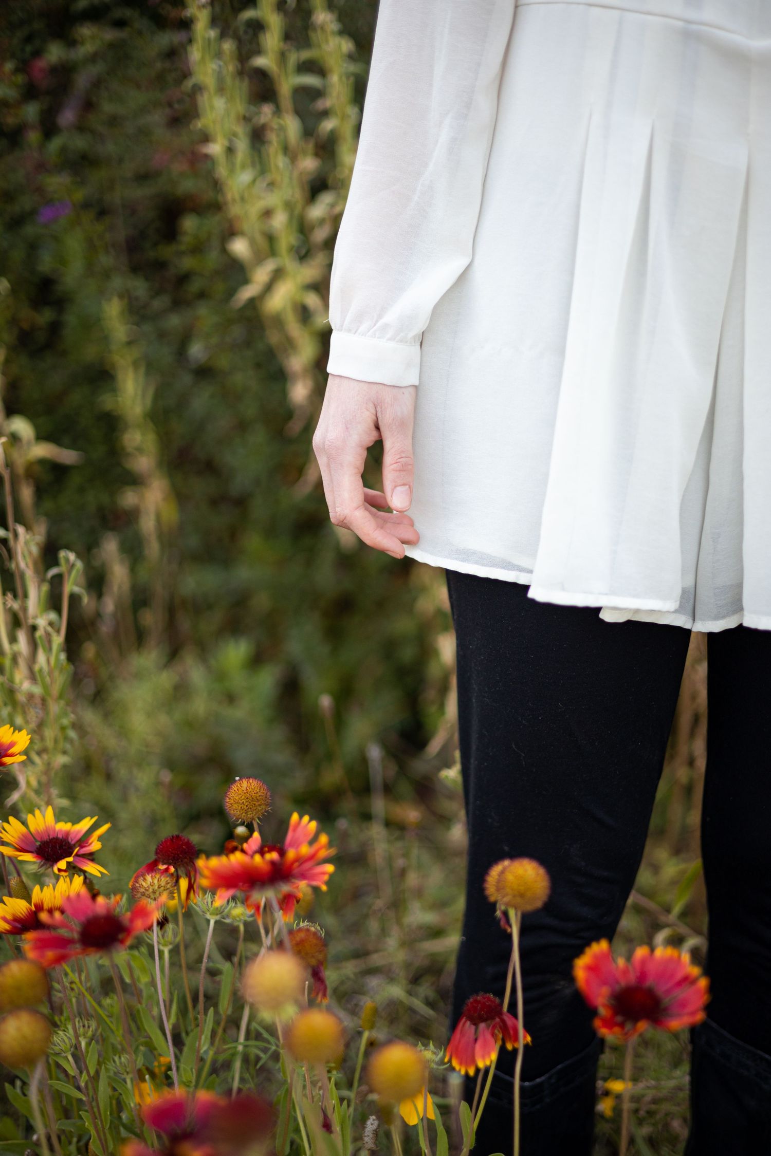 A person in a white flowing shirt and black leggings stands among blooming red and orange Blanket flowers in a garden.