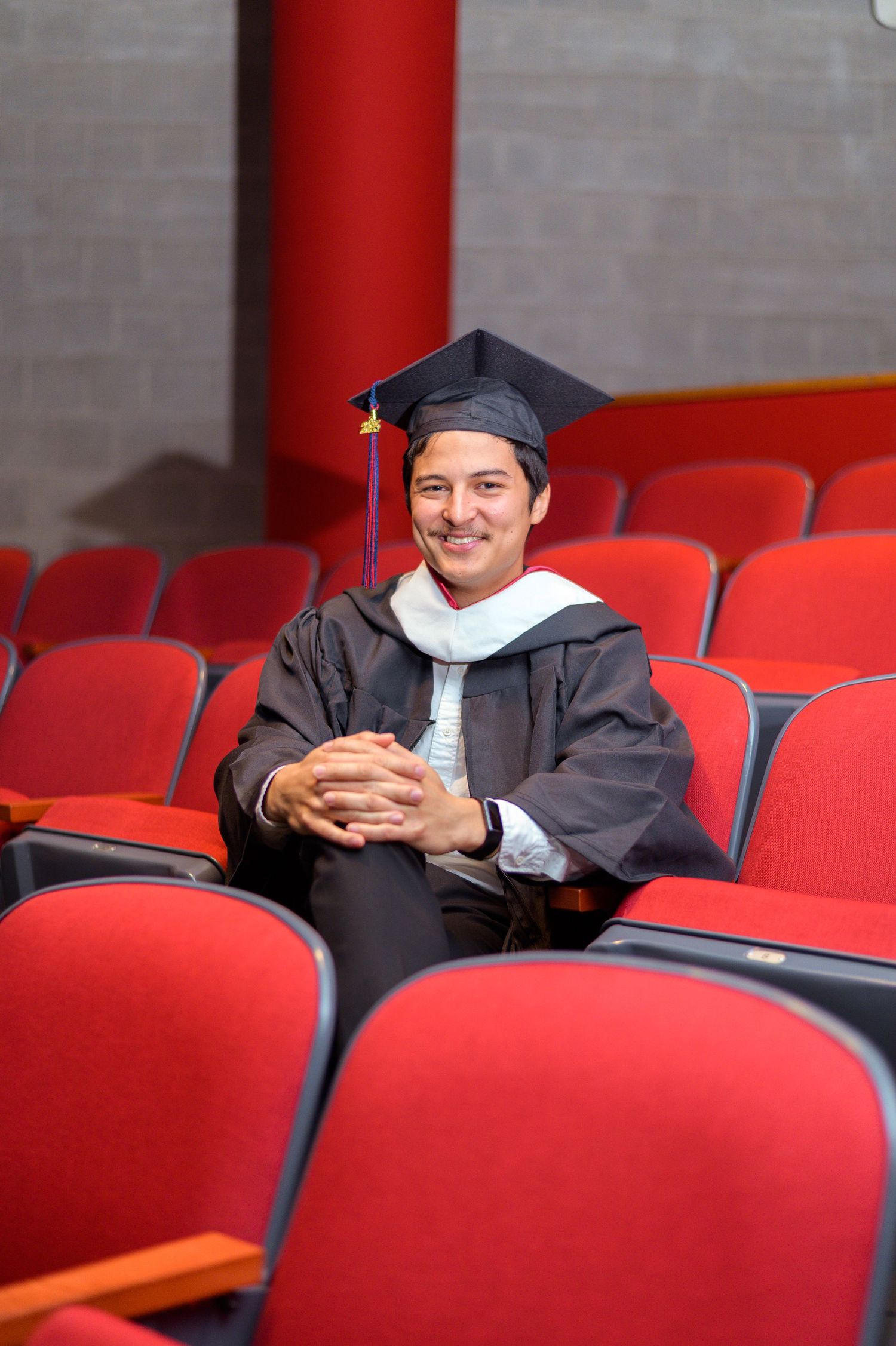 Graduate in traditional cap and gown sitting in red auditorium seating.