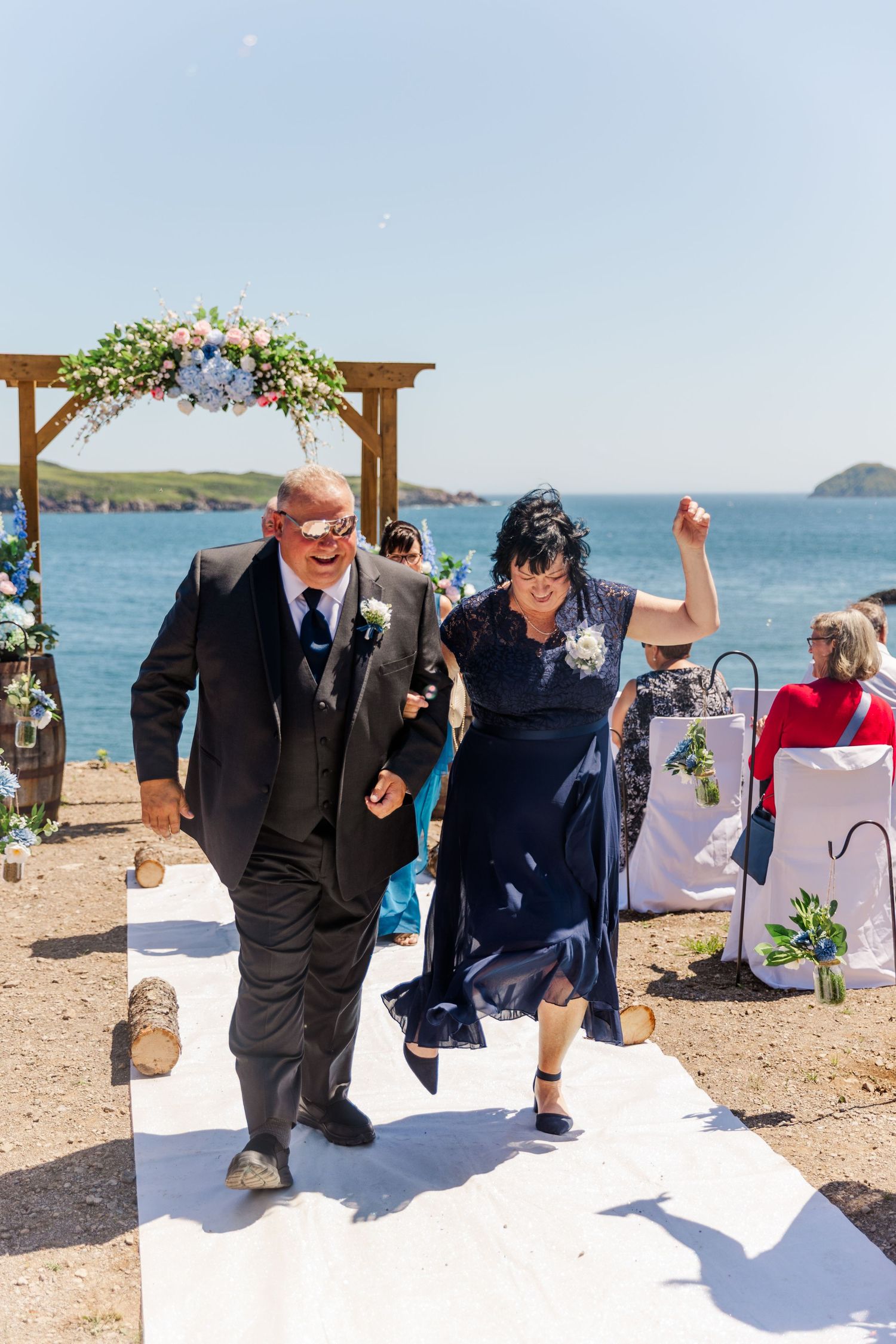 A couple joyfully walks down a white aisle runner at an outdoor oceanside wedding ceremony under a floral-decorated arch.