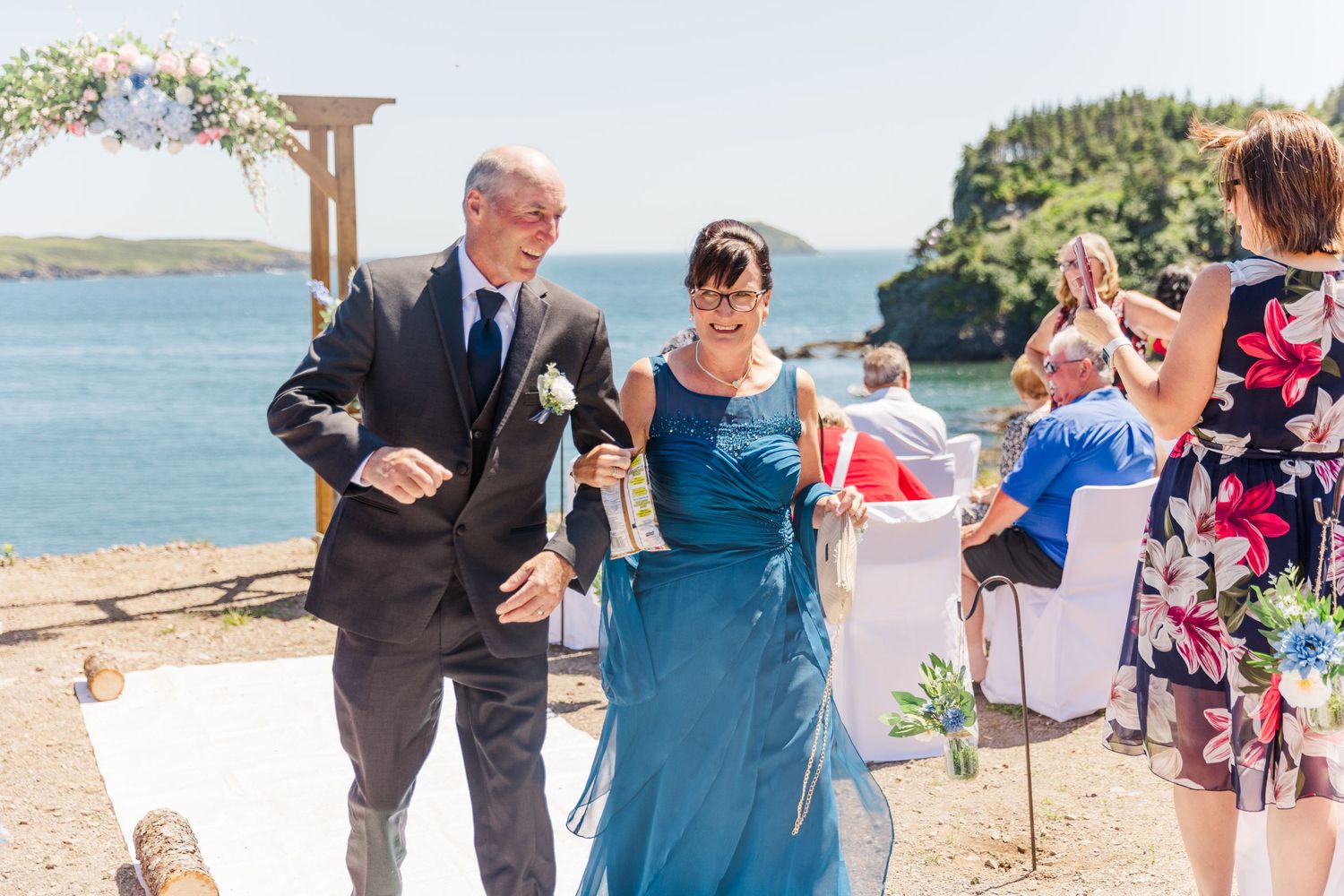 A couple celebrates in blue attire under a wooden arch by the ocean during an outdoor wedding ceremony.