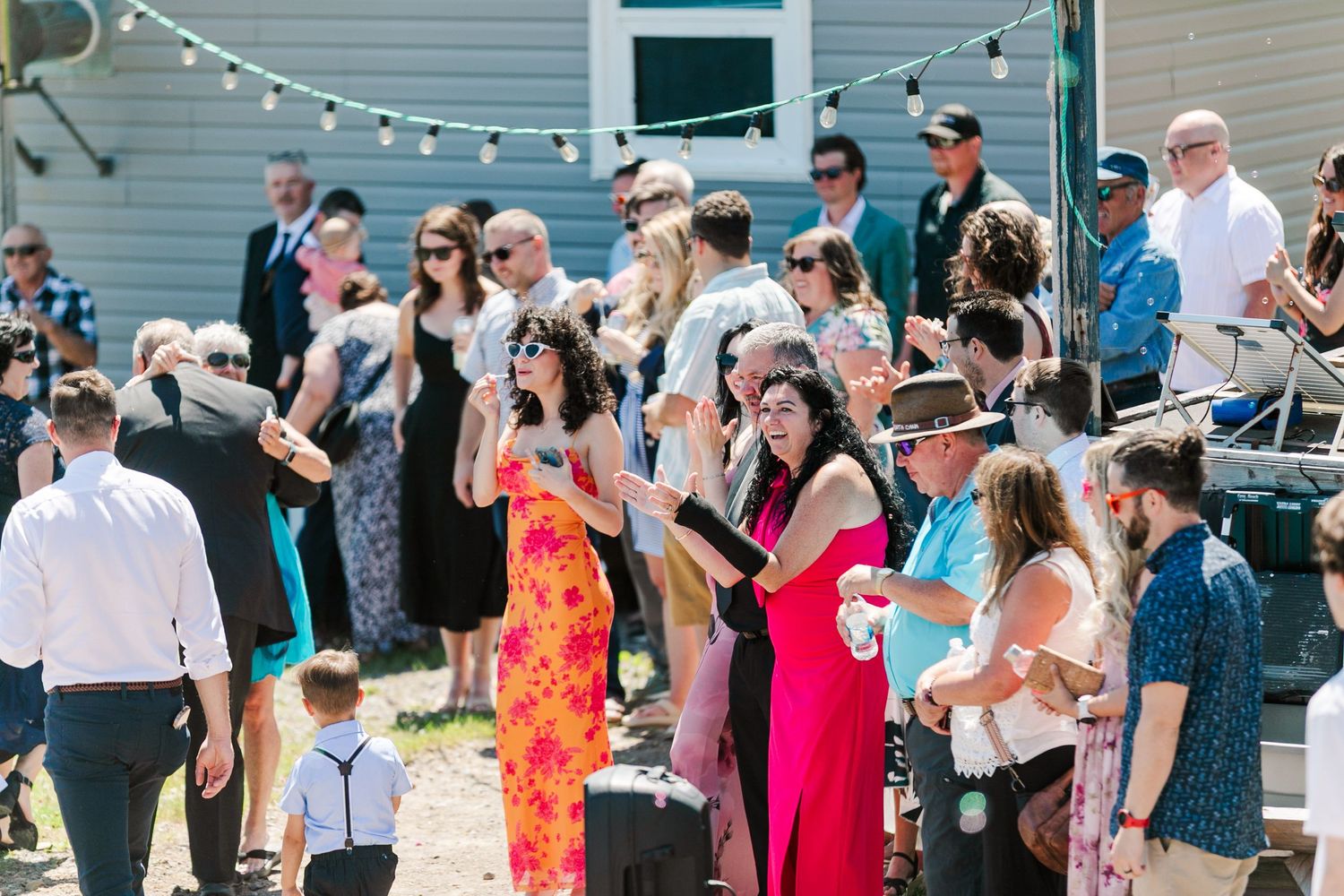 A large group mingles and socializes at an outdoor party with string lights overhead on a summer day.