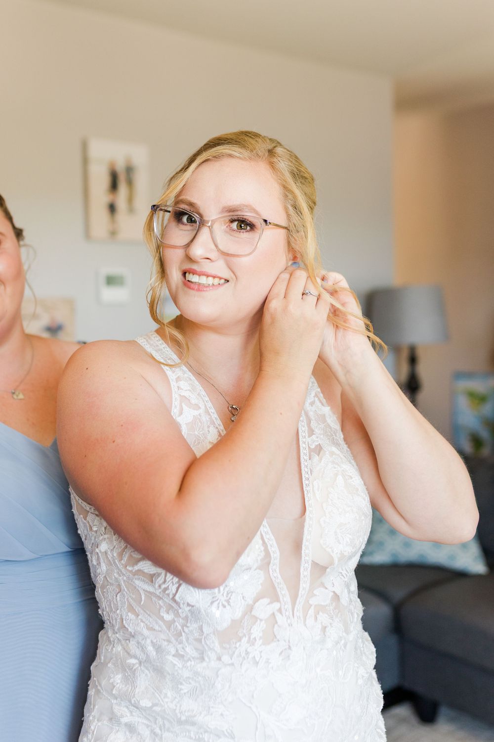 A bride in a white lace wedding dress smiles with joy while getting ready for her wedding day in a bright room.